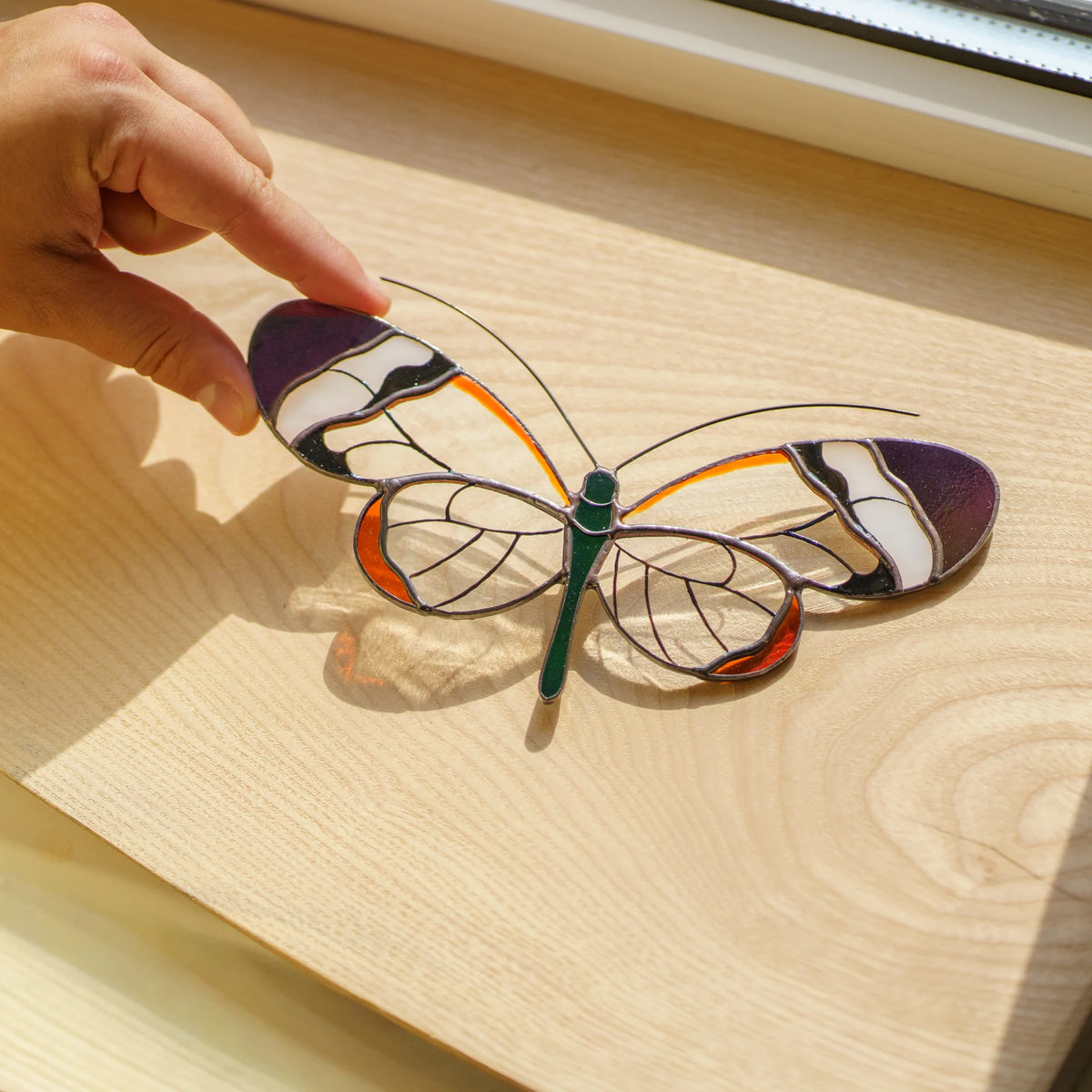 Glasswing butterfly suncatcher resting on a wooden windowsill, clear wings throwing soft color onto the grain