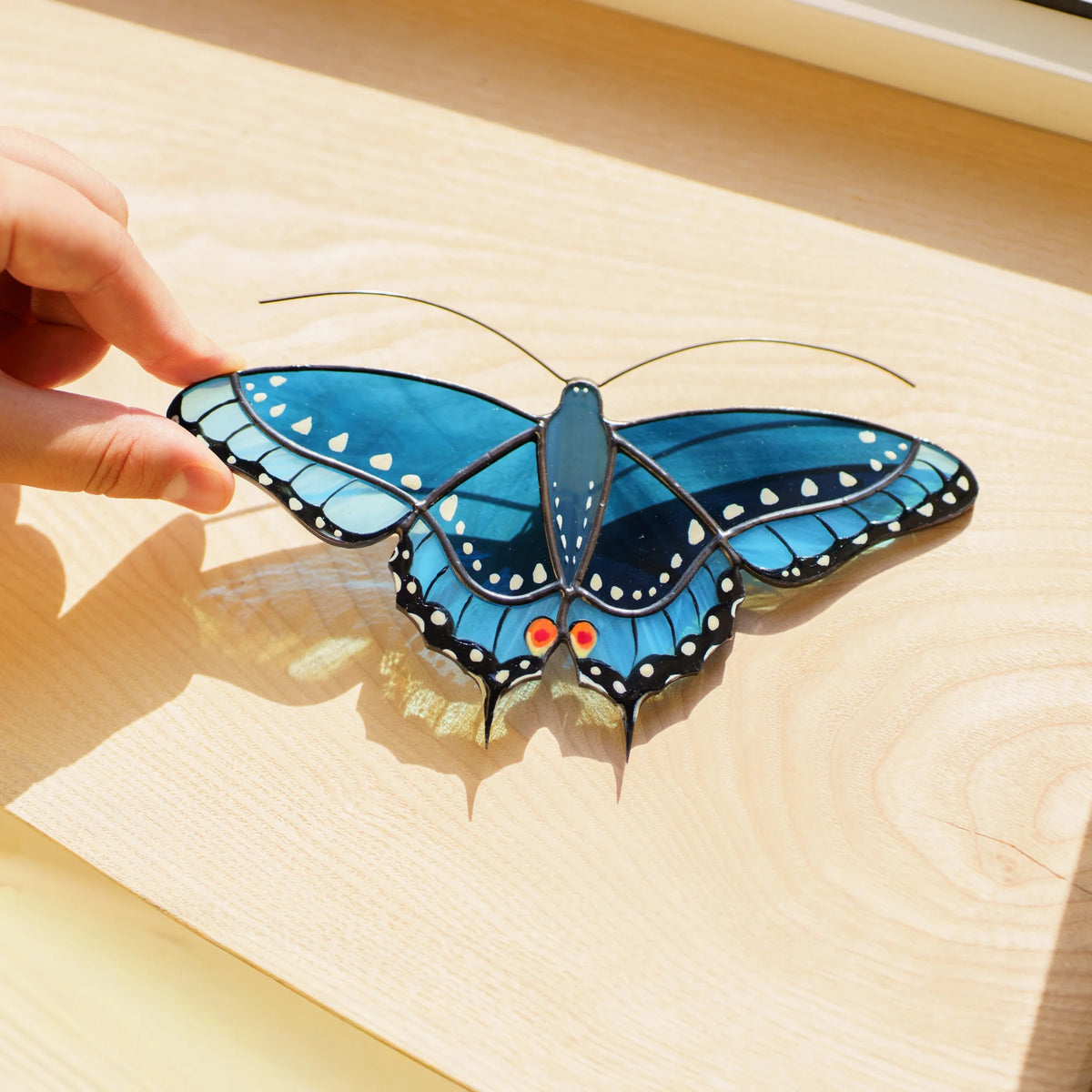 Blue swallowtail stained glass suncatcher on a wooden sill, sunlight passing through its blue glass