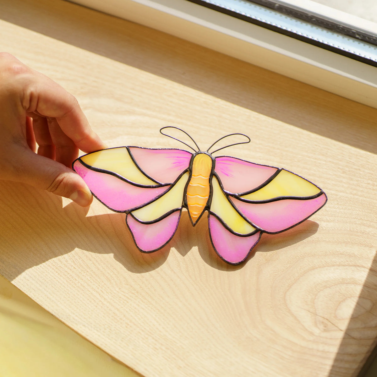 Rosy maple moth suncatcher in pink and yellow glass resting on a warm wooden windowsill