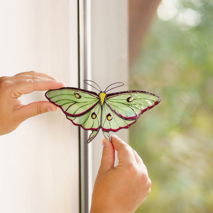 Luna moth stained glass suncatcher held against a window frame, green wings in front of a leafy view
