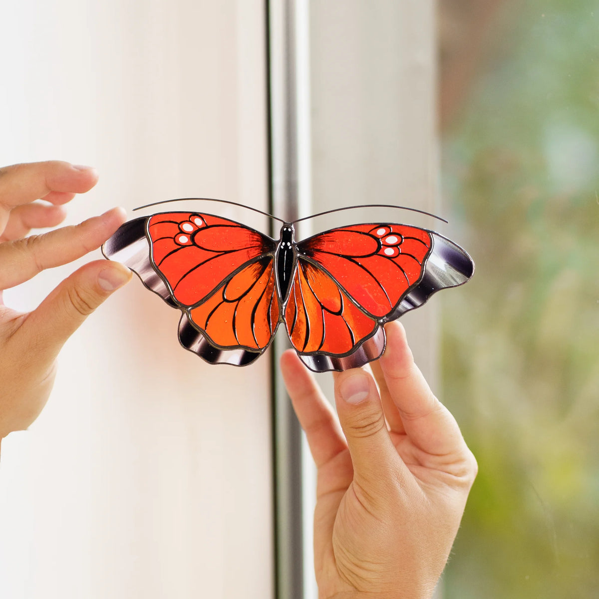 Orange butterfly suncatcher held up beside a white window frame, bright glass against a soft garden view