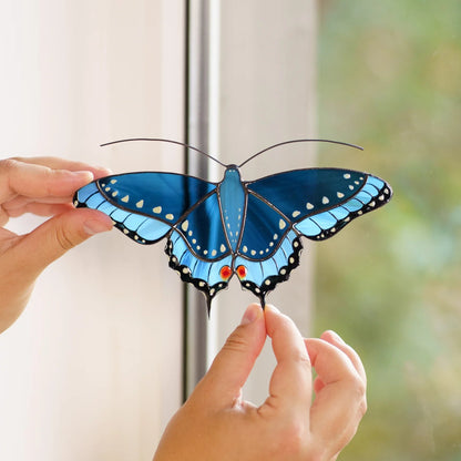 Hands placing a blue swallowtail stained glass suncatcher on a window, fingertip touching one tail
