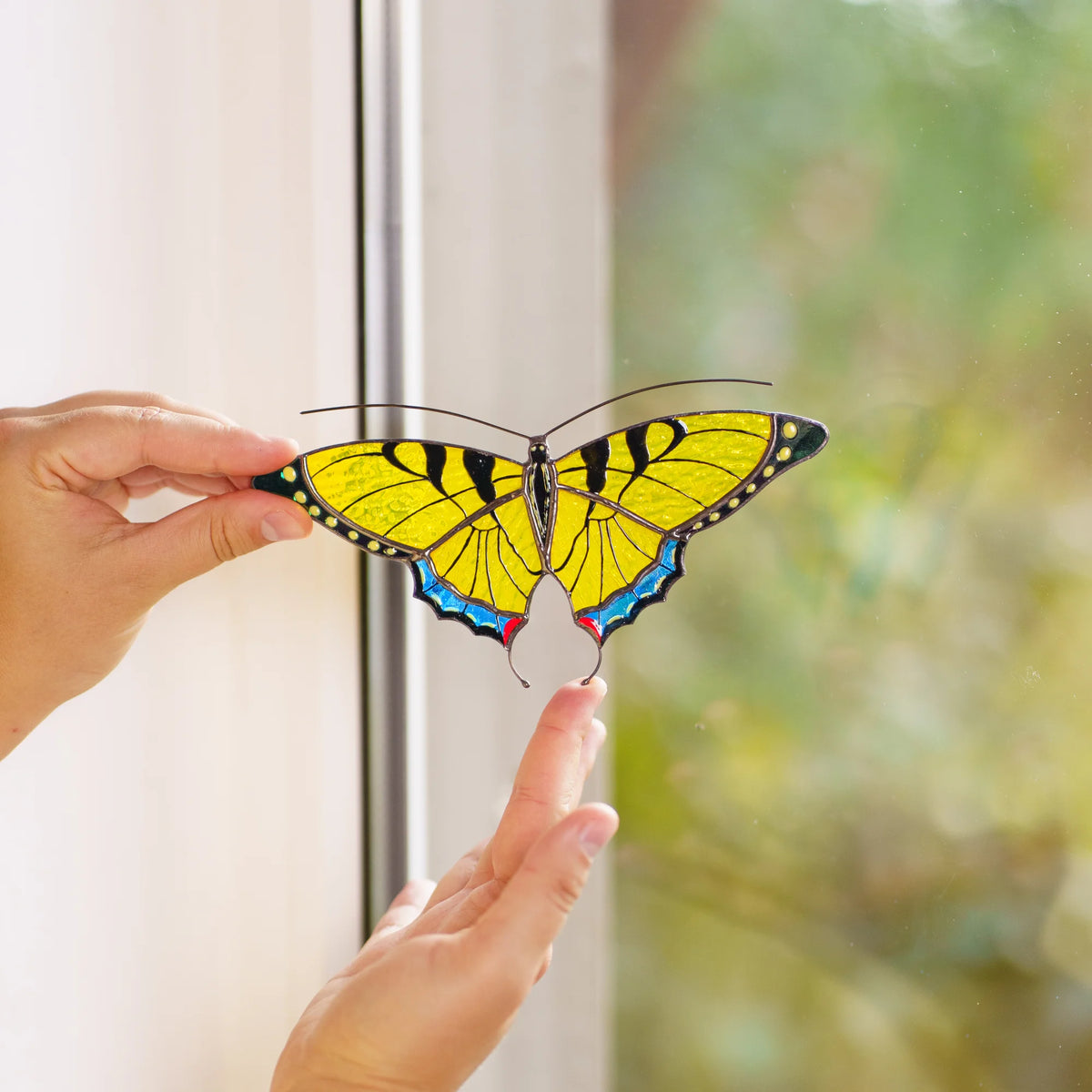Yellow swallowtail suncatcher held against a window frame, hands framing its curled tails