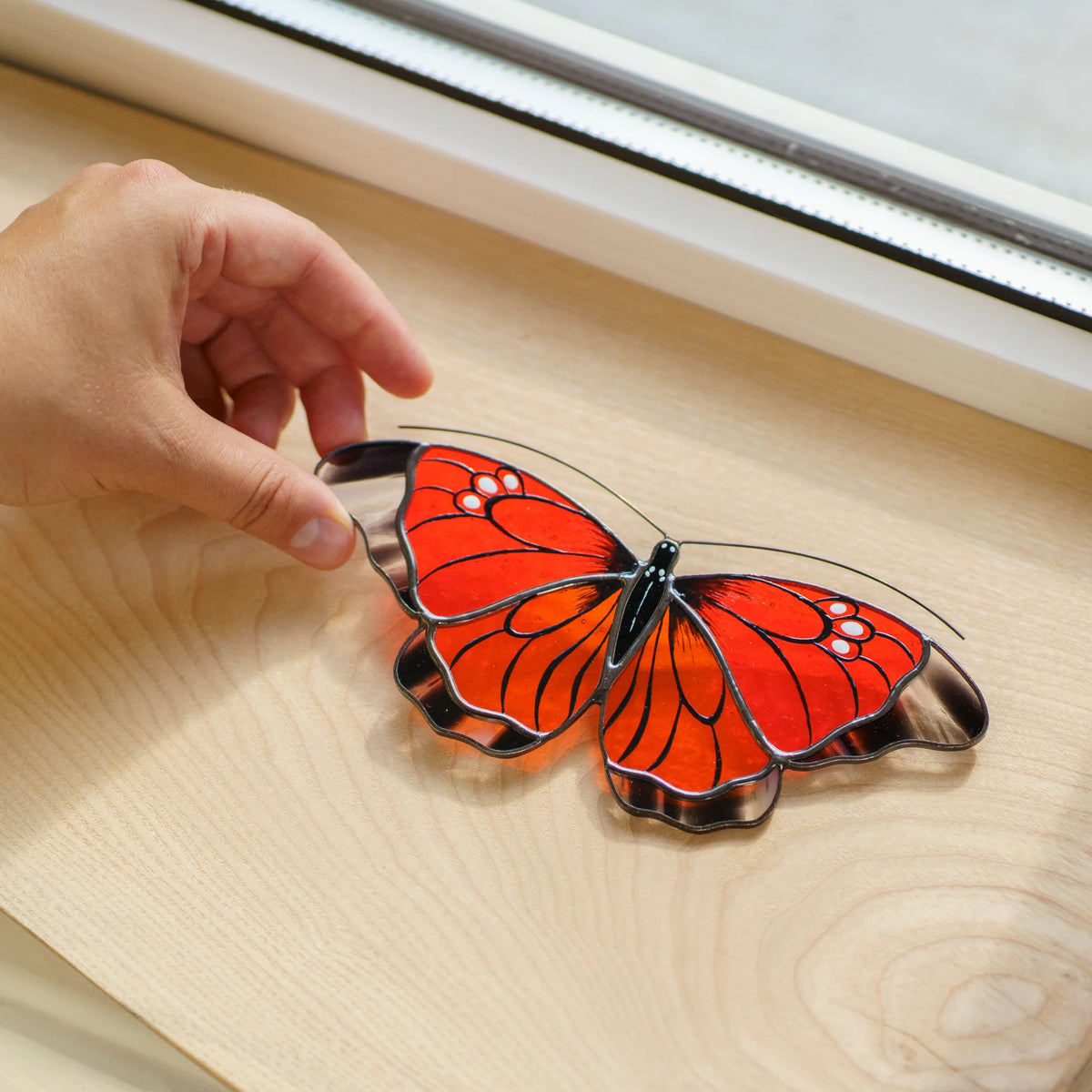 Orange butterfly suncatcher on a wooden windowsill, a hand lifting its clear glass wingtip
