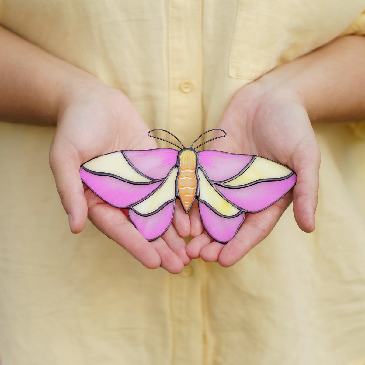 Rosy maple moth suncatcher in pink and cream glass held in open palms against a yellow shirt
