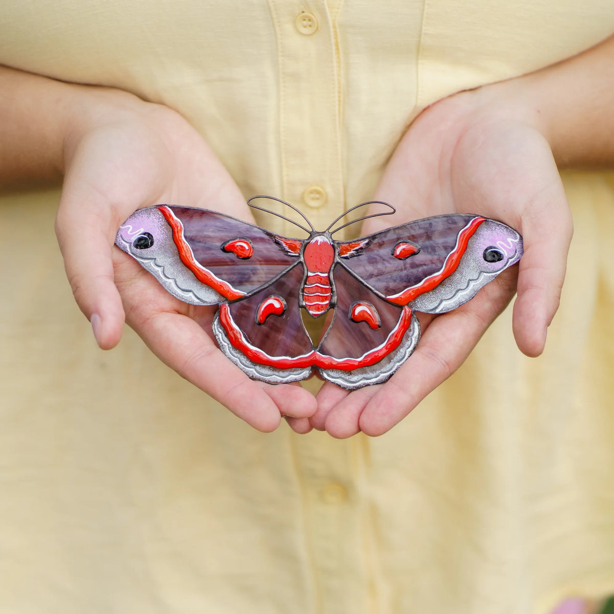 Cecropia moth stained glass suncatcher in plum and red glass with white eyespots held in cupped hands