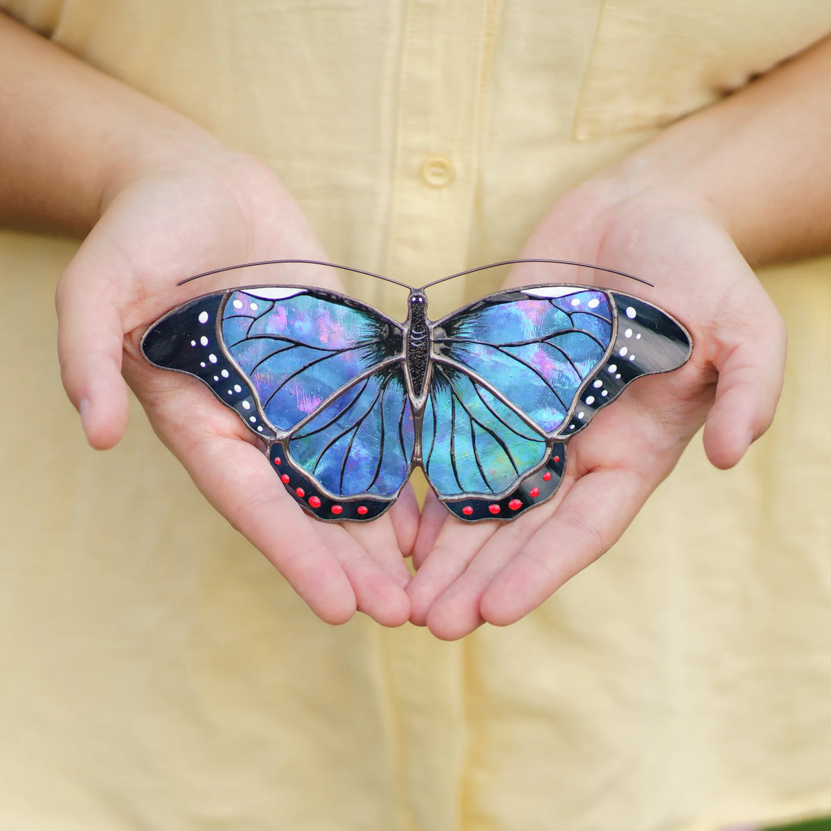Blue morpho suncatcher in iridescent blue and violet glass with red dots held in cupped hands