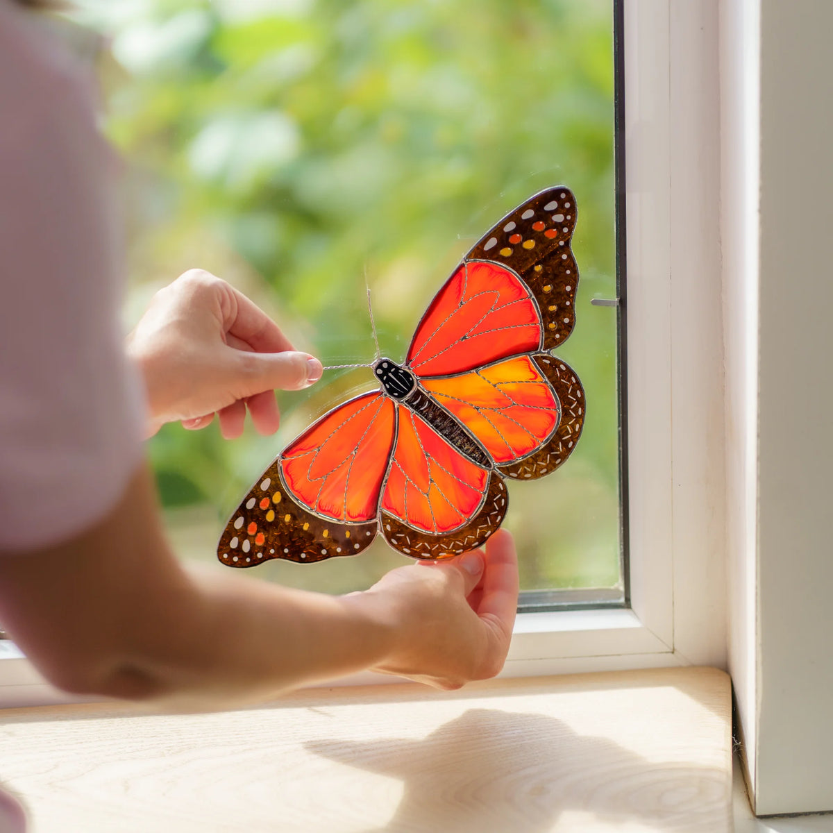 Hands pressing the orange monarch into a window corner, a small metal clip visible on the frame
