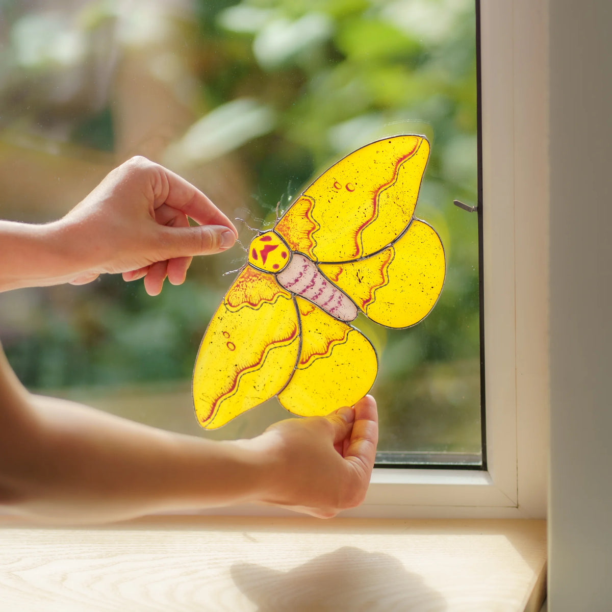 Hands fitting a yellow moth suncatcher into the corner of a window with a small metal clip