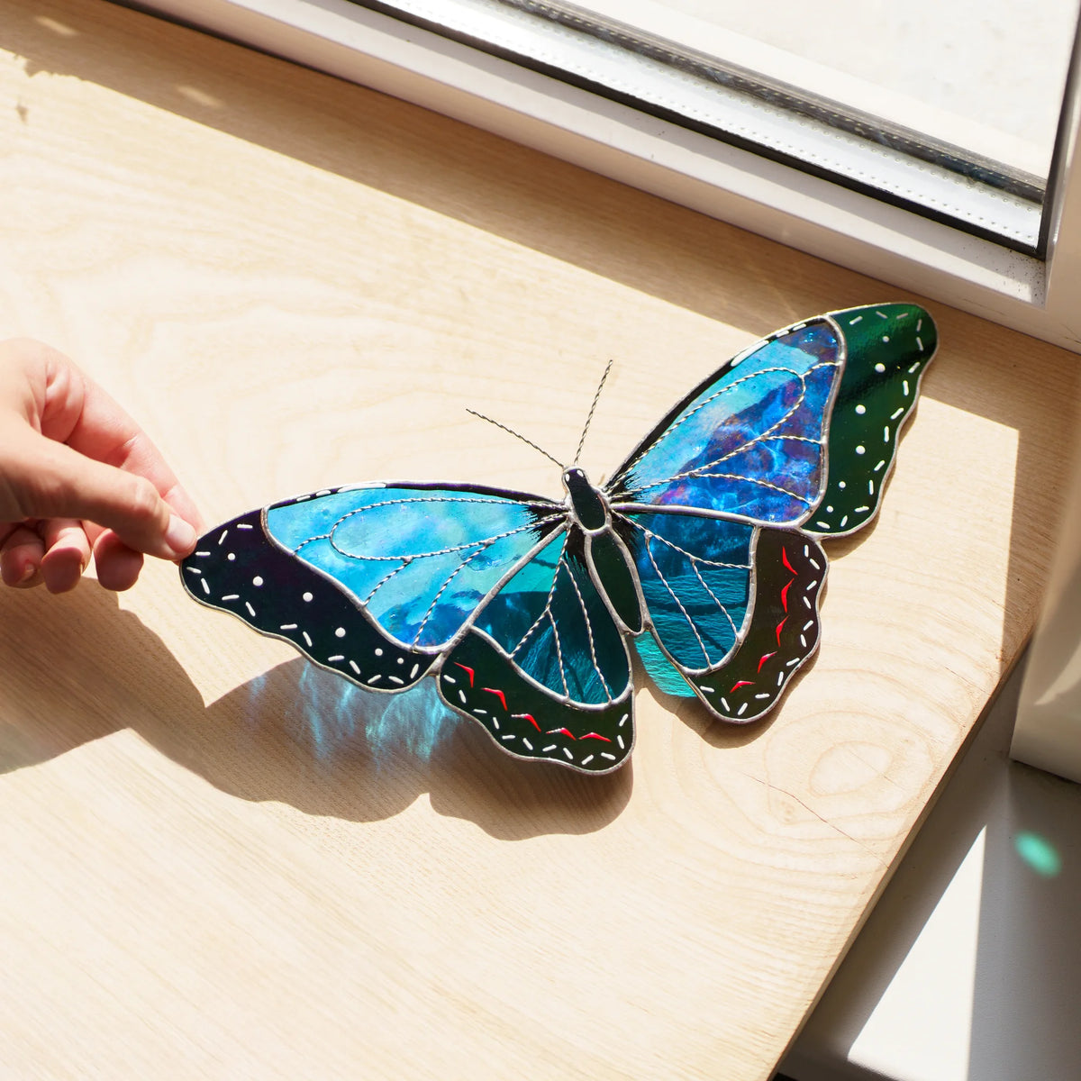 Morpho suncatcher on a sunlit wooden sill, shimmering blue glass with red and white dash details