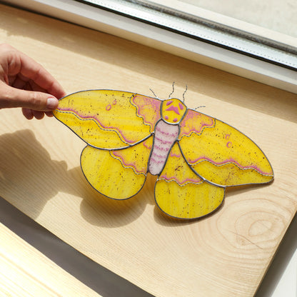 Imperial moth glass art on a wooden windowsill, pink wing bands and speckled yellow glass in the sun