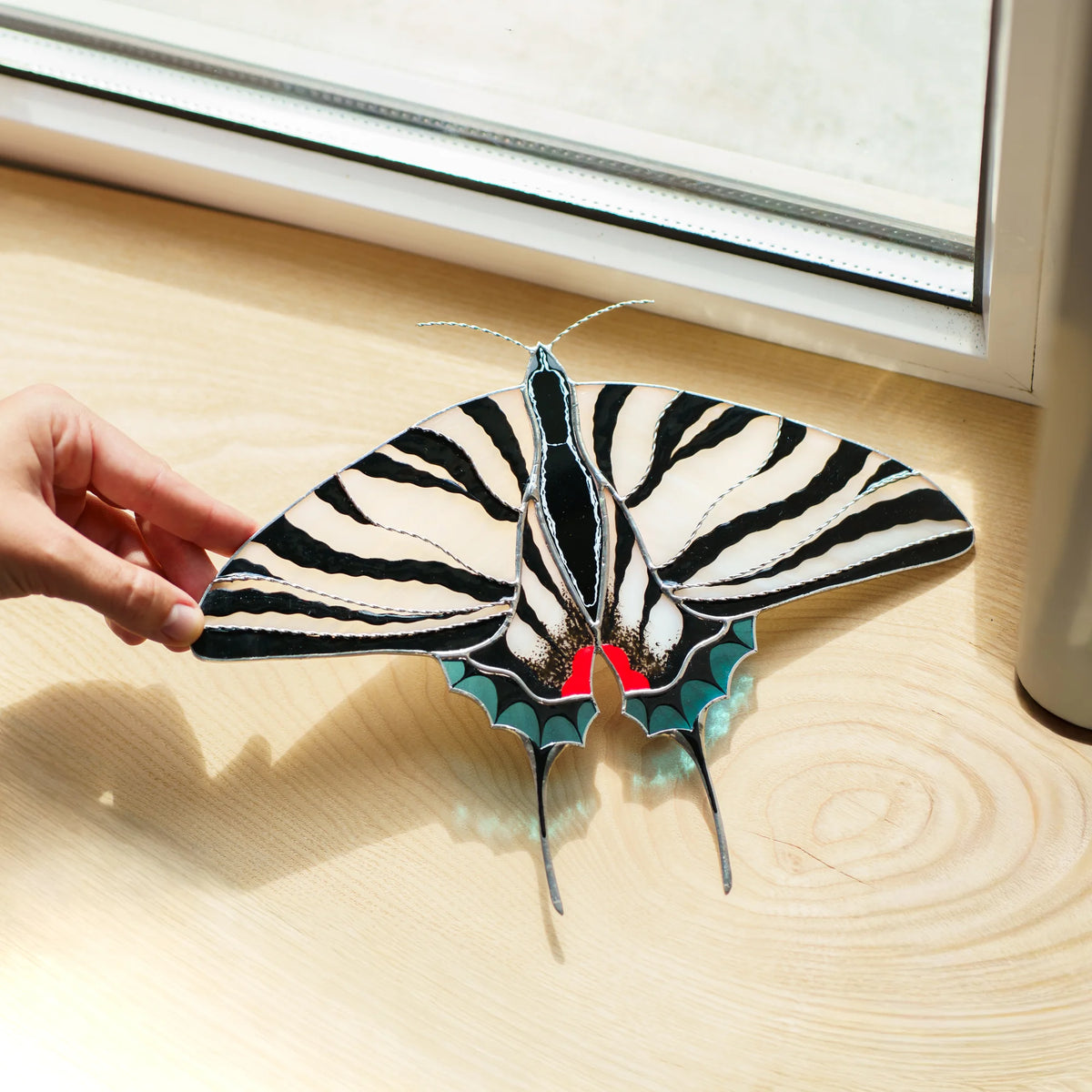Zebra swallowtail suncatcher laid flat on a wooden sill, red spots and teal edges glowing in the sun