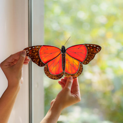 Two hands holding the orange and amber butterfly to a bright window, garden blurred behind