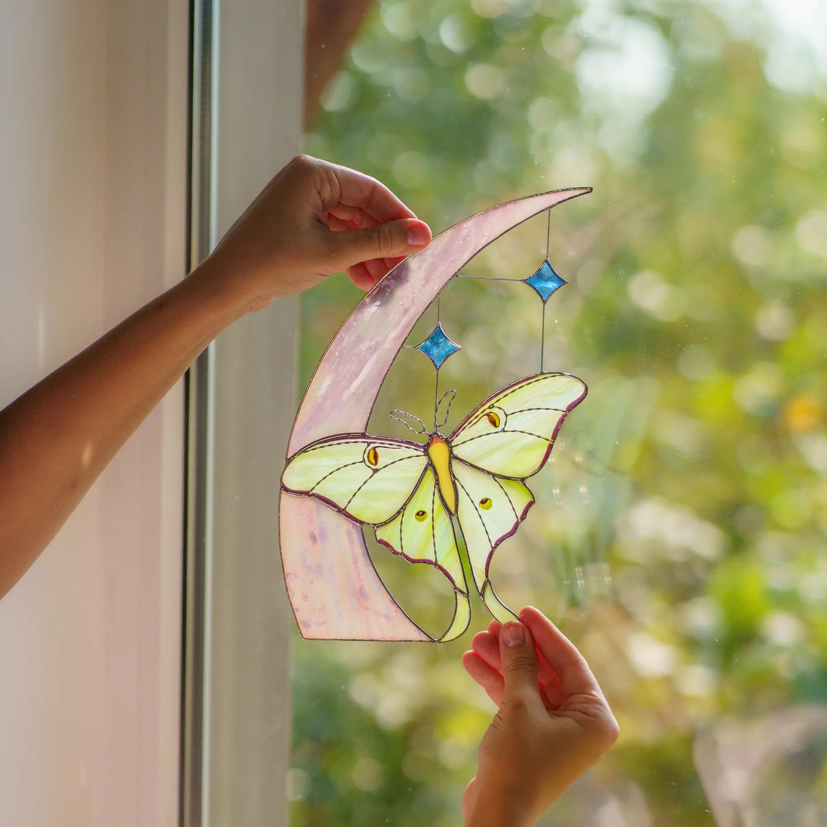 Two hands holding the moon and moth suncatcher to a bright window, garden blurred behind