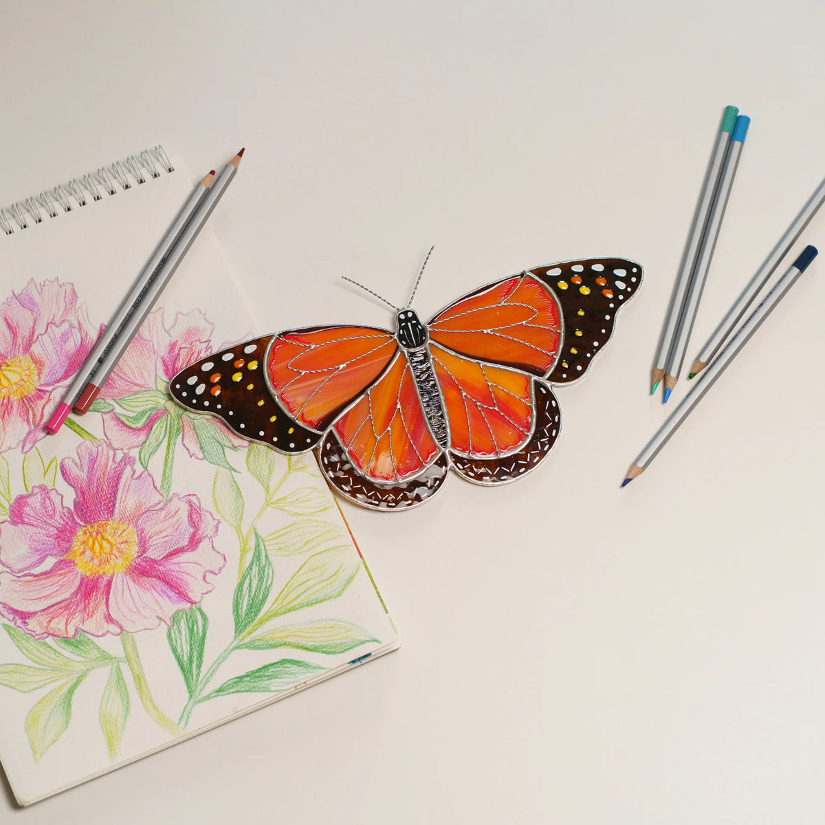 Monarch stained glass on a white table beside a pink peony drawing and colored pencils