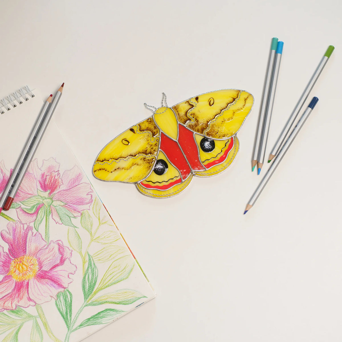 Io moth stained glass on a white table beside a pink peony drawing and colored pencils