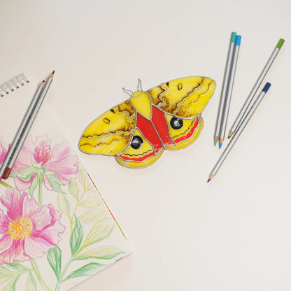 Io moth stained glass on a white table beside a pink peony drawing and colored pencils