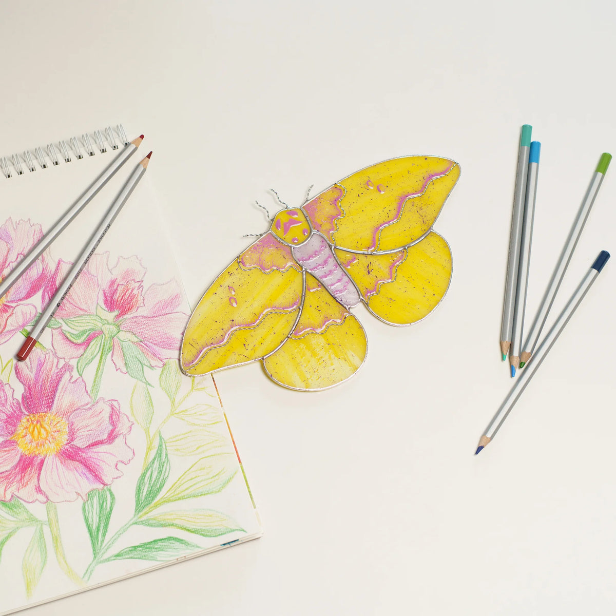 Imperial moth suncatcher beside a sketchbook with a pink peony drawing and colored pencils