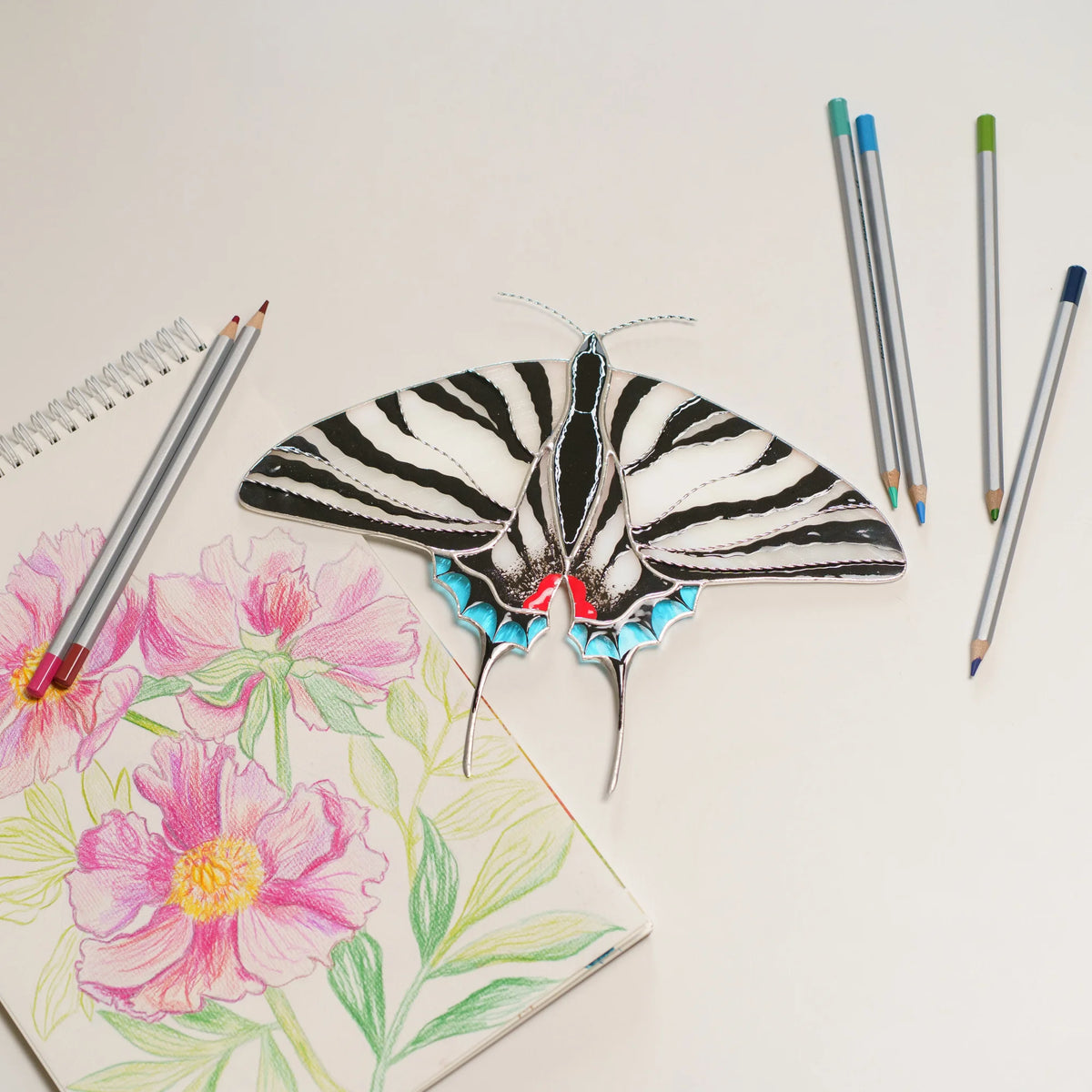 Zebra swallowtail stained glass beside a peony drawing and colored pencils on a white table