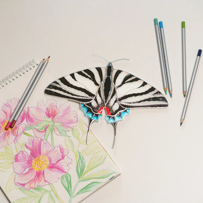 Zebra swallowtail stained glass beside a peony drawing and colored pencils on a white table