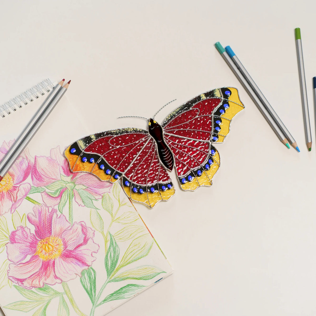 Mourning cloak suncatcher on a white desk next to a peony sketch and colored pencils