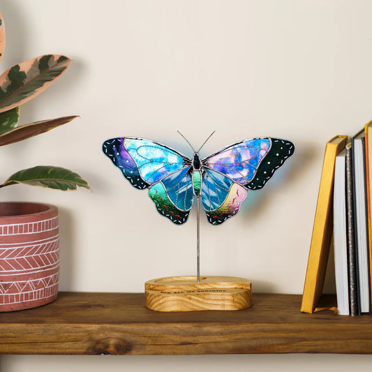 Blue morpho butterfly stained glass suncatcher on its wooden stand beside potted plants and books