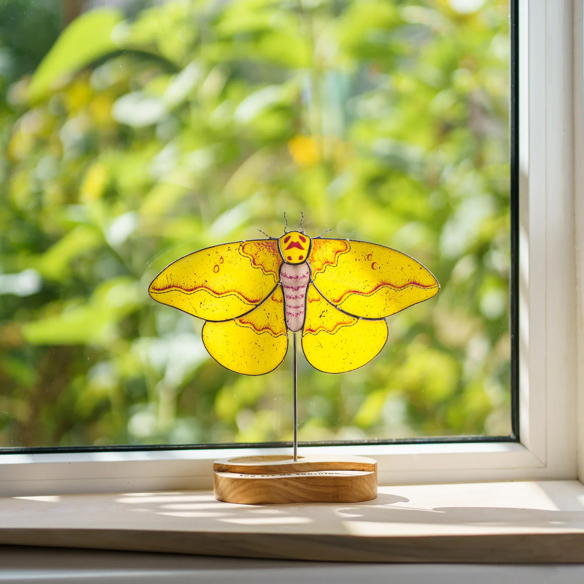 Stained glass imperial moth suncatcher on a windowsill with a garden view behind it