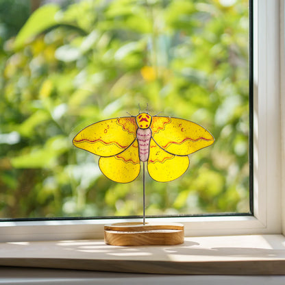 Stained glass imperial moth suncatcher on a windowsill with a garden view behind it