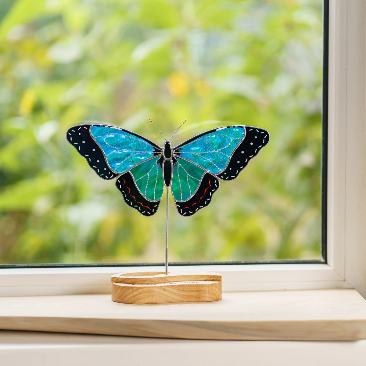 Stained glass blue morpho butterfly suncatcher on a windowsill with a garden view behind it