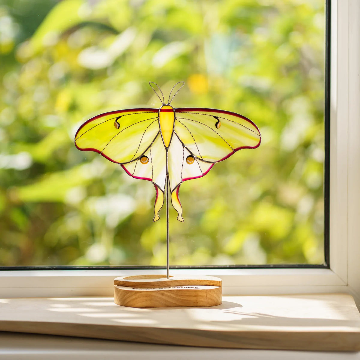 Stained glass luna moth suncatcher glowing in sunlight on a windowsill with a garden view