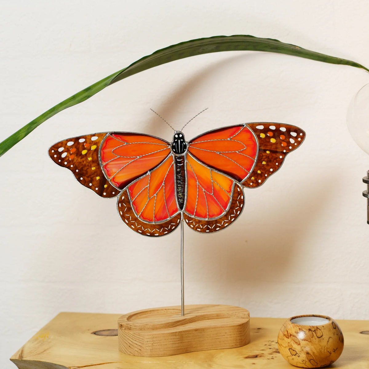 Monarch butterfly stained glass suncatcher on its wooden stand beside a green leaf