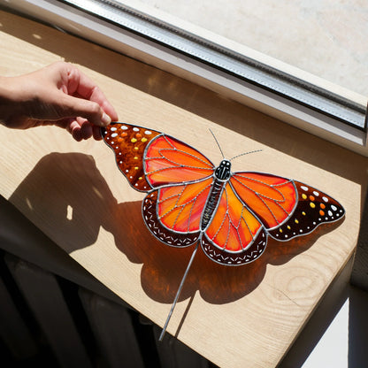 Hand holding the wing of a stained glass monarch butterfly suncatcher on a sunlit windowsill