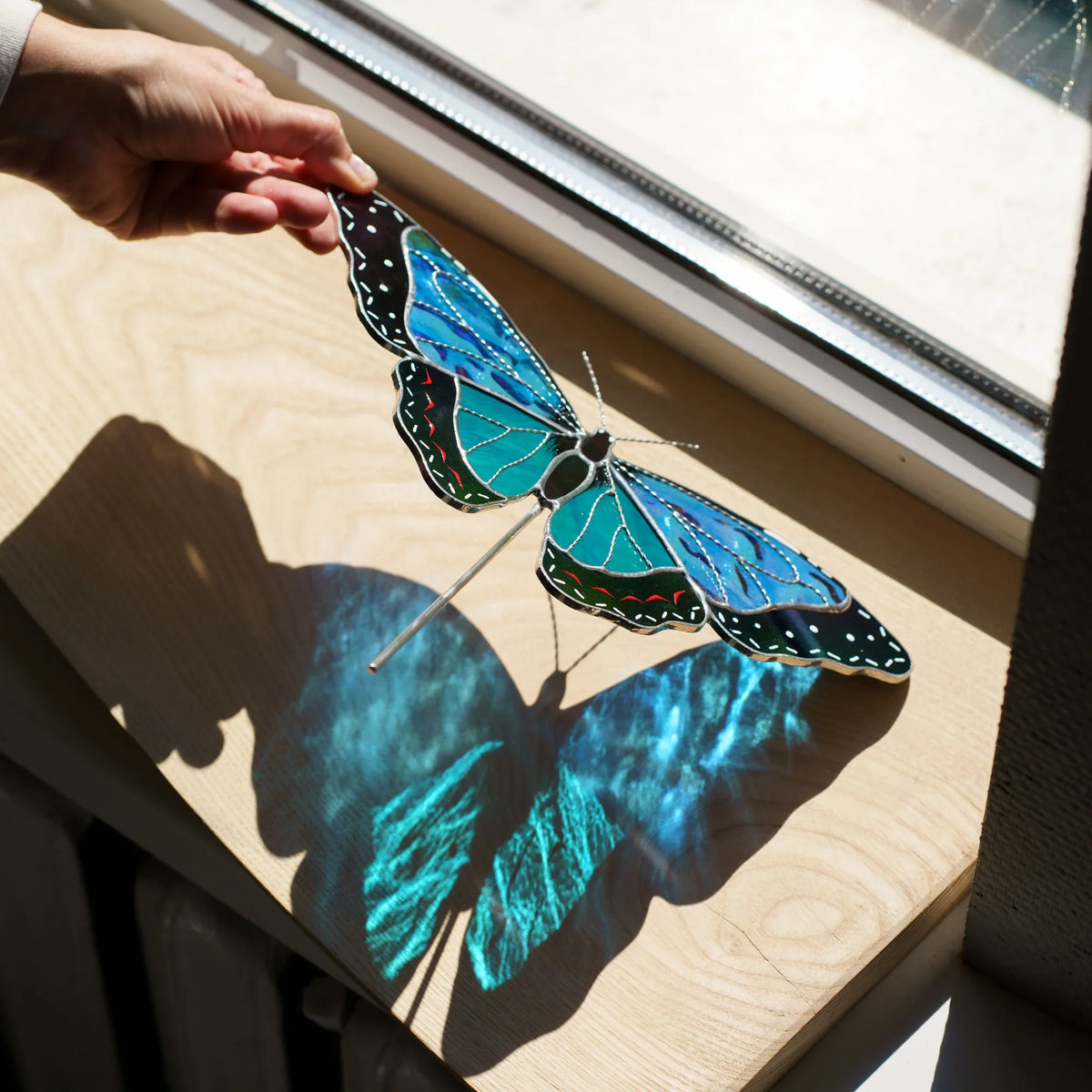 Hand holding the wing of a stained glass blue morpho suncatcher, casting a blue shadow