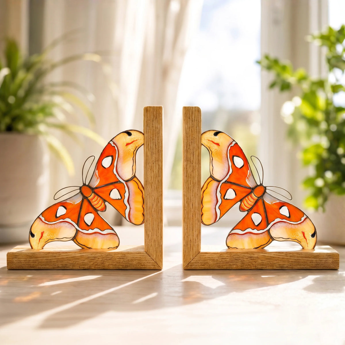 A pair of vibrant orange and yellow stained glass moth bookends sitting on a wooden surface against a bright windowsill, perfect as a unique and practical gift.