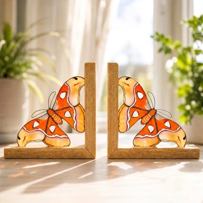 A pair of vibrant orange and yellow stained glass moth bookends sitting on a wooden surface against a bright windowsill, perfect as a unique and practical gift.