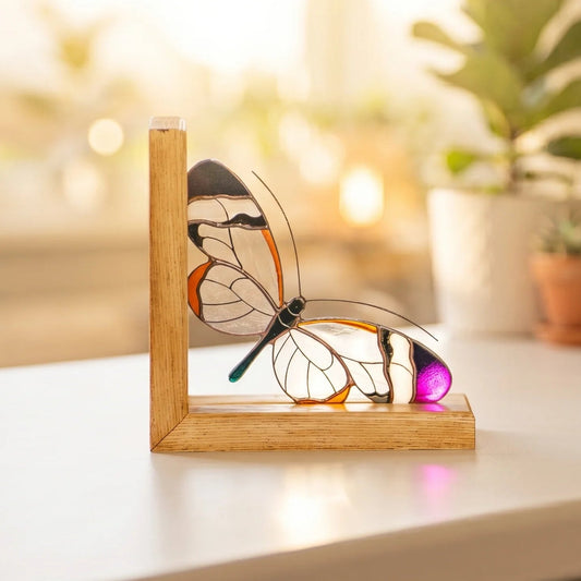 A stained glass glasswing butterfly sculpture resting on a light wooden bookend, placed on a white windowsill with a softly blurred background of potted plants and warm sunlight.
