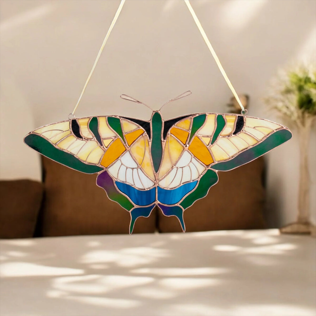 A stained-glass window hanging butterfly ornament with multicolored panels in green, blue, yellow, white, and purple, suspended by thin cords in a softly lit indoor setting.