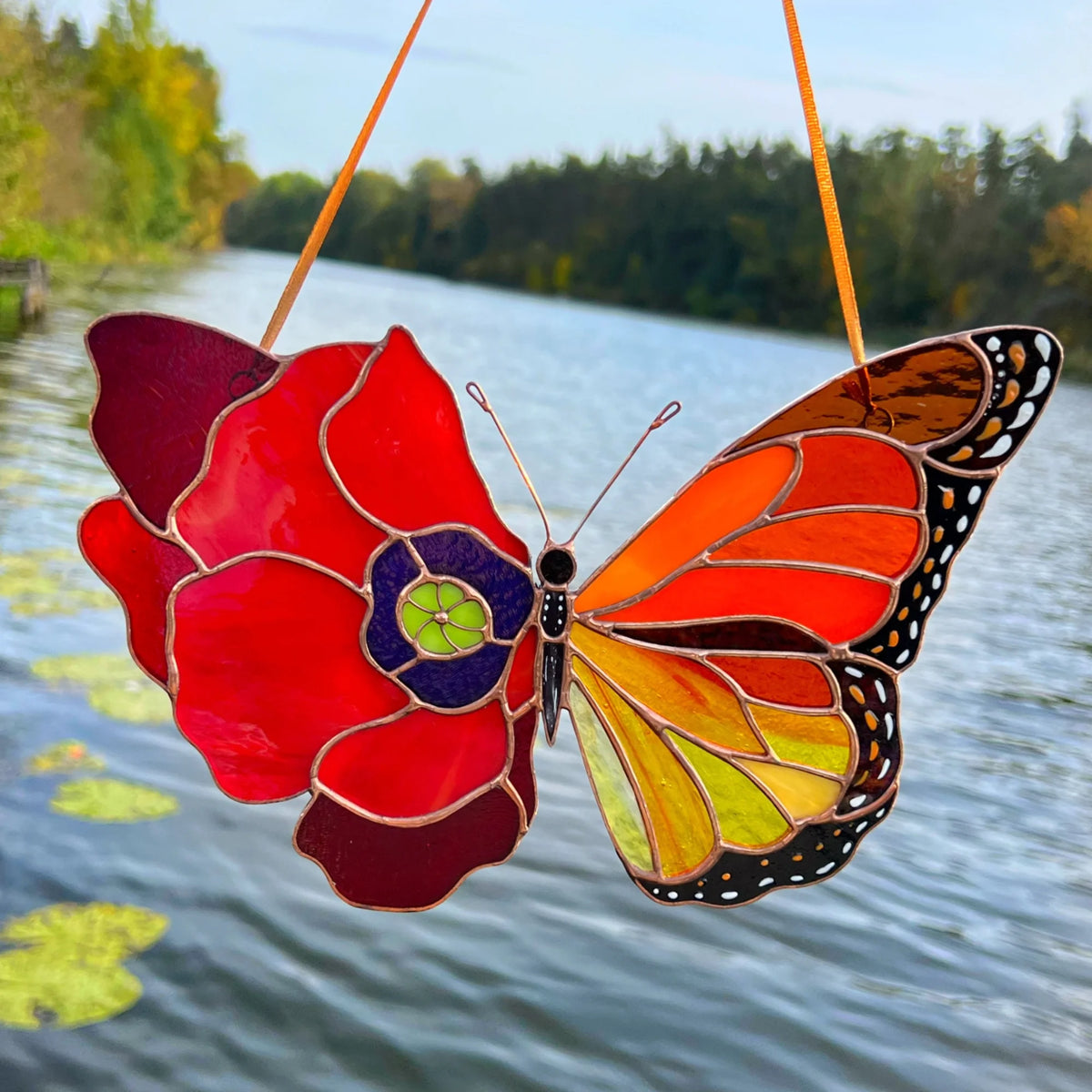 This exquisite stained glass poppy-monarch butterfly hangs outdoors, capturing the natural light with a scenic river and forest in the background.