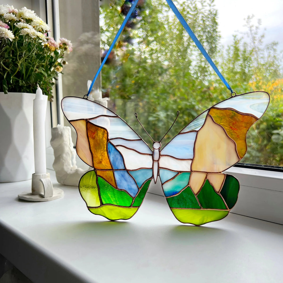 A stained glass butterfly suncatcher with a blue hanging ribbon, displayed on a white windowsill. The butterfly features a Yosemite landscape design with green hills, blue sky, and warm yellow and orange clouds, outlined in dark brown lead lines.