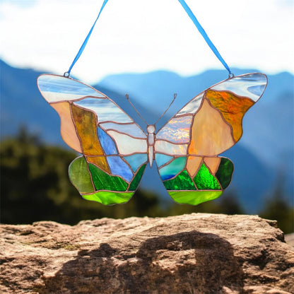 A stained glass butterfly suncatcher with a blue hanging ribbon, suspended outdoors in front of a blurred Yosemite mountain background. The butterfly features a multicolor landscape design with blue, green, orange, and beige sections outlined in gold-toned metal.