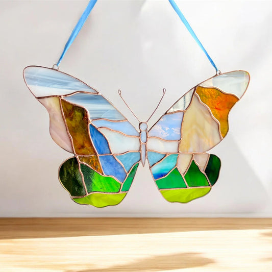 A stained glass butterfly suncatcher with a Yosemite landscape design, hanging from a blue ribbon against a white wall with sunlight casting a shadow on a wooden surface below.