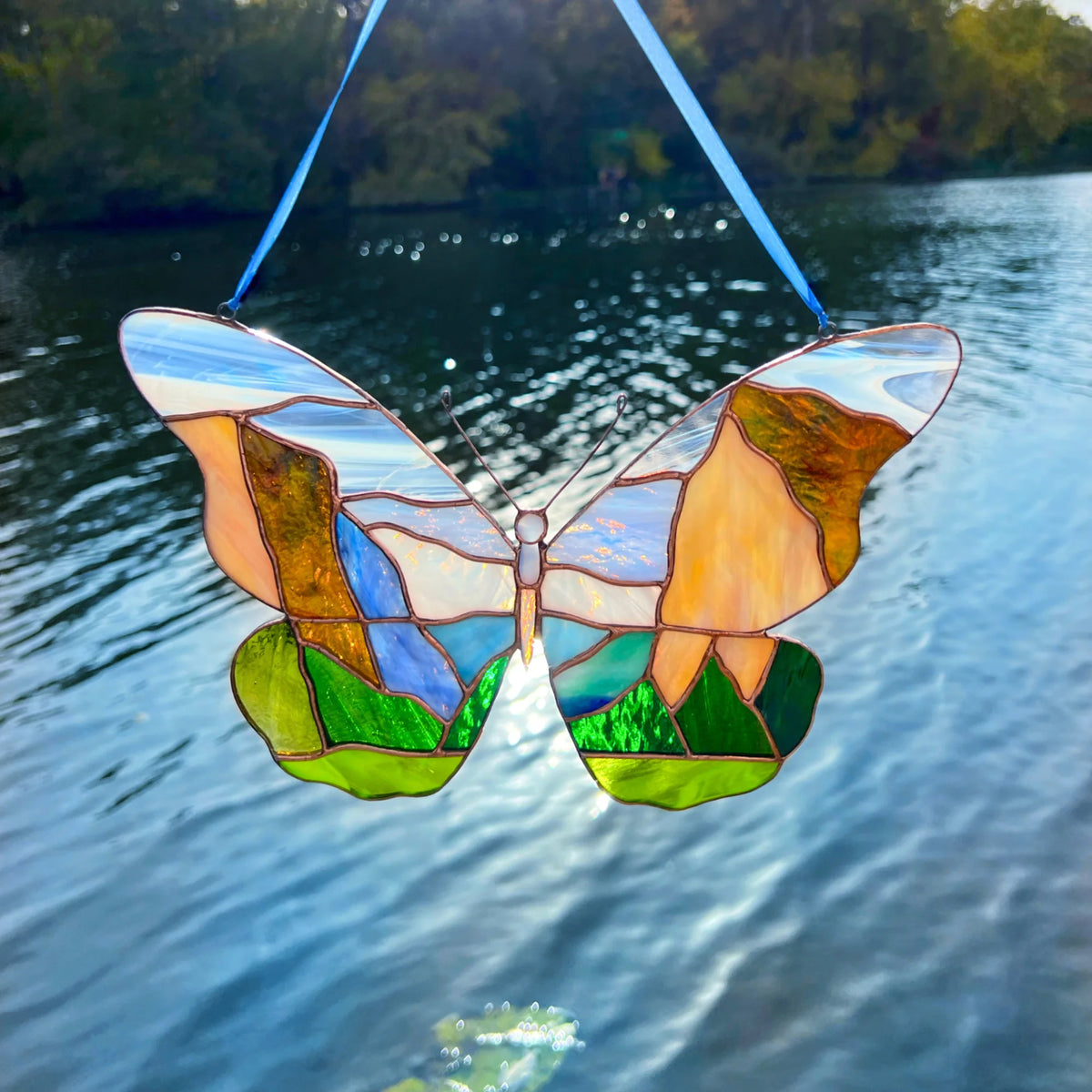 A stained glass butterfly suncatcher with Yosemite landscape, suspended by blue ribbons against a background of rippling blue water and a tree-lined shore.