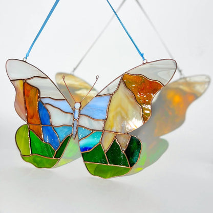 A stained glass butterfly suncatcher with a colorful Yosemite landscape design, featuring blue sky, green hills, and orange and yellow sections, suspended by a blue string against a white background.