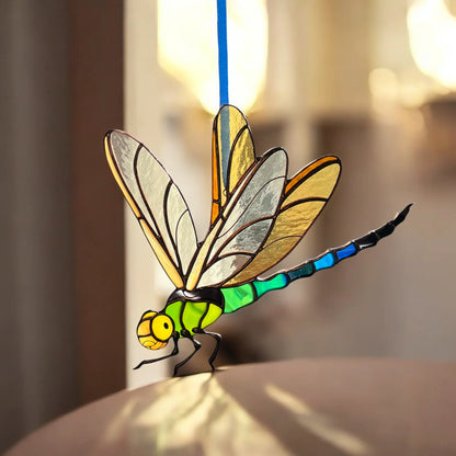 A stained-glass dragonfly ornament with a blue hanging string, featuring yellow, green, and blue segments, displayed on a reflective surface with a warm, blurred background.