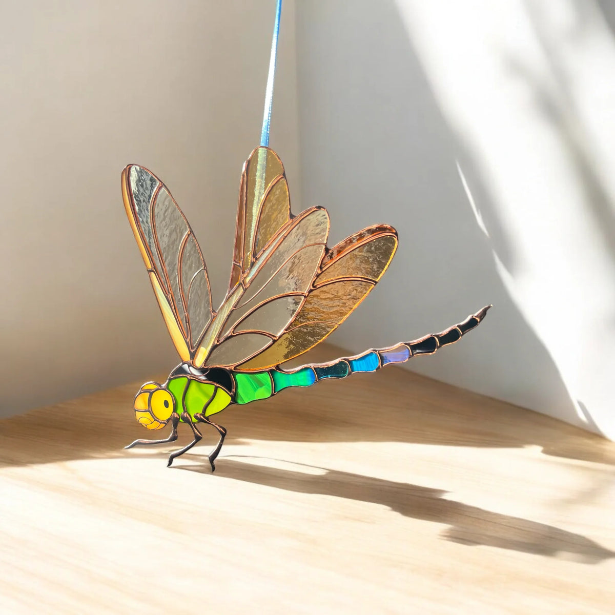 A stained glass dragonfly suncatcher with a bright green and blue segmented body, yellow head, and golden-brown wings, displayed on a light wooden surface against a white wall with soft shadows.