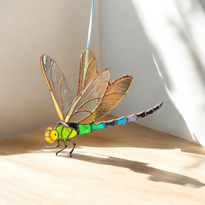 A stained glass dragonfly suncatcher with a bright green and blue segmented body, yellow head, and golden-brown wings, displayed on a light wooden surface against a white wall with soft shadows.