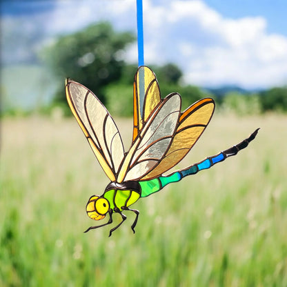 A stained-glass dragonfly ornament with yellow, green, blue, black, and translucent wings, suspended by a blue string against a blurred outdoor field and sky background.