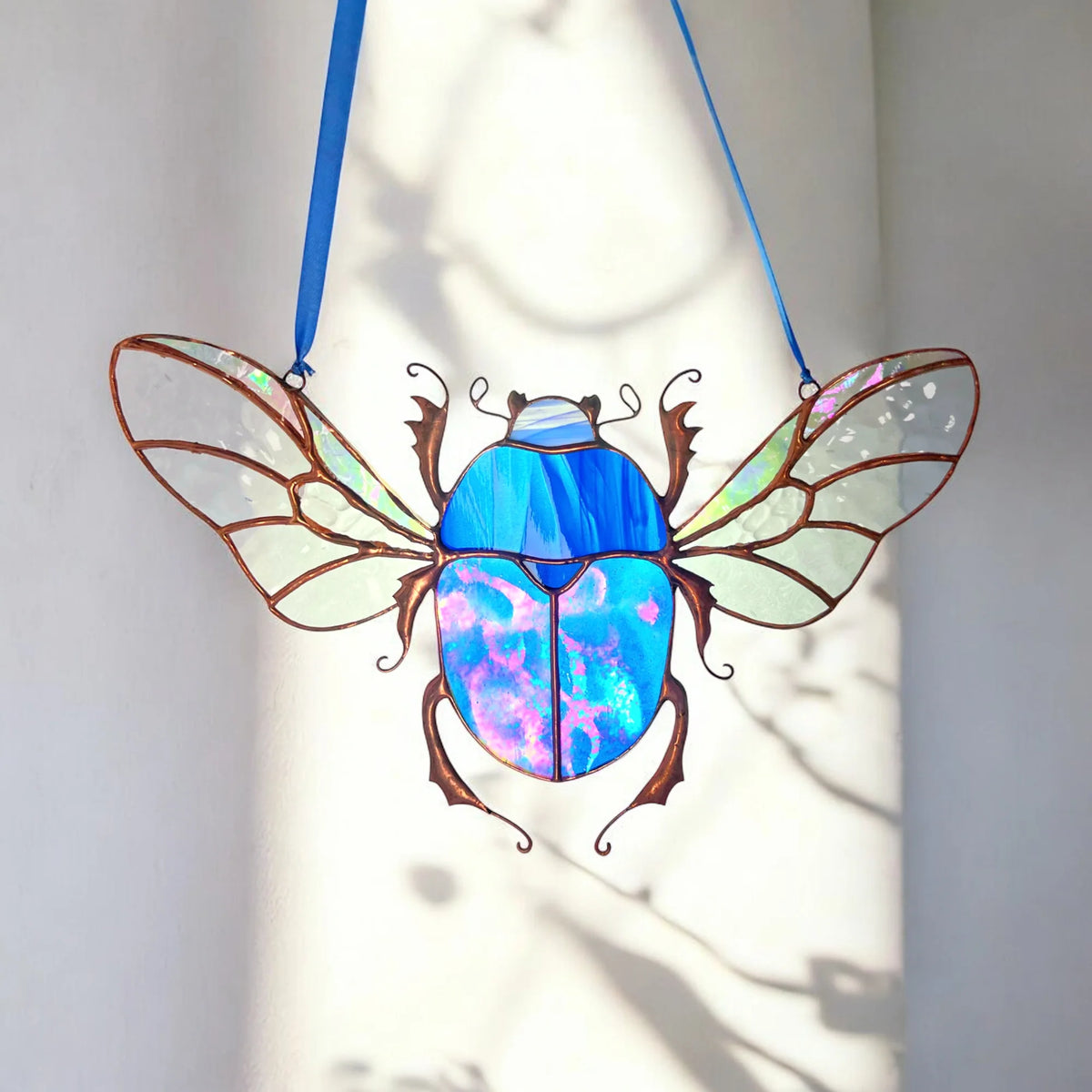 A stained glass beetle suncatcher with iridescent blue and purple wings, suspended by two blue ribbons against a white wall with soft shadow patterns.