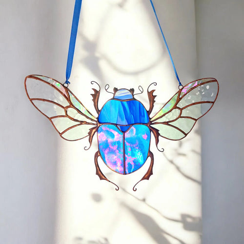 A stained glass beetle suncatcher with iridescent blue and purple wings, suspended by two blue ribbons against a white wall with soft shadow patterns.
