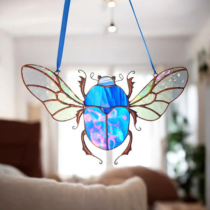 A stained glass beetle ornament with iridescent blue and purple wings and body, suspended by two blue ribbons in a softly blurred living room setting.